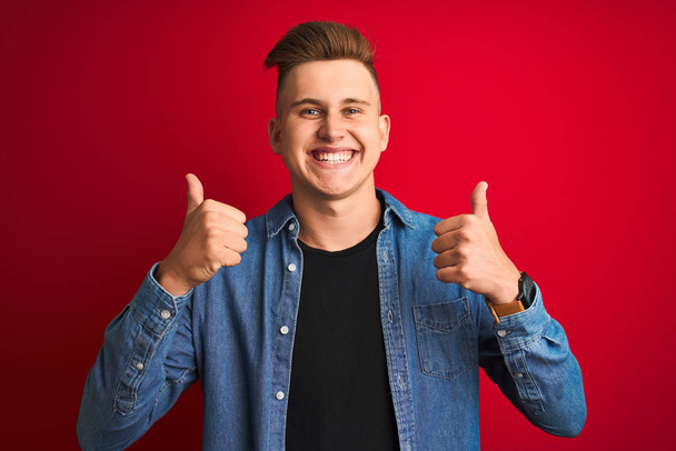 Ein junger gutaussehender Mann in Jeanshemd steht über einem vereinzelten roten Erfolgsschild und macht eine positive Geste mit der Hand, Daumen hoch lächelnd und glücklich. Fröhlicher Ausdruck und Siegergeste. - Foto, Bild