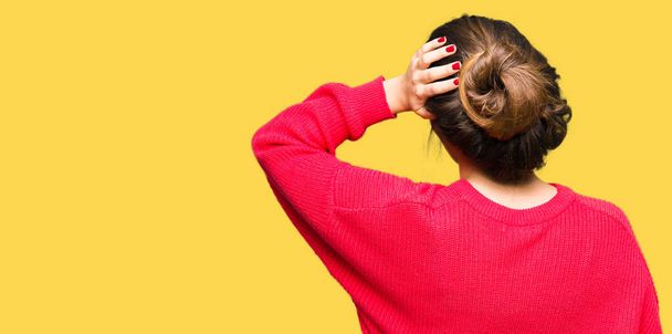 Young beautiful woman wearing red sweater and bun Backwards thinking about doubt with hand on head - Photo, Image