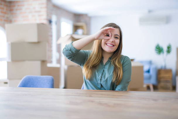 Mujer joven sentada en la mesa con cajas de cartón detrás de su mudanza a un nuevo hogar haciendo un buen gesto con la mano sonriente, los ojos mirando a través de los dedos con la cara feliz
. - Foto, imagen