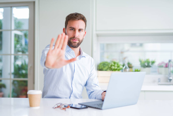 Красивий чоловік працює за допомогою комп'ютерного ноутбука і п'є чашку кави з відкритою рукою робить знак зупинки з серйозним і впевненим виразом, захисний жест
 - Фото, зображення