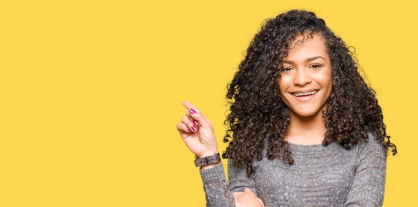 Young beautiful woman with curly hair wearing grey sweater with a big smile on face, pointing with hand and finger to the side looking at the camera. - Photo, image