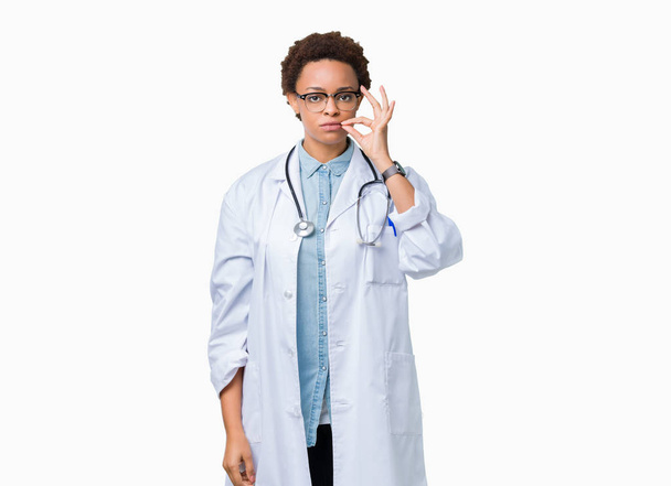 Młody lekarz african american kobieta sobie medycznych płaszcz nad na białym tle usta i usta jako zip palcami. Sekret i niema, Tabu, rozmowa - Zdjęcie, obraz