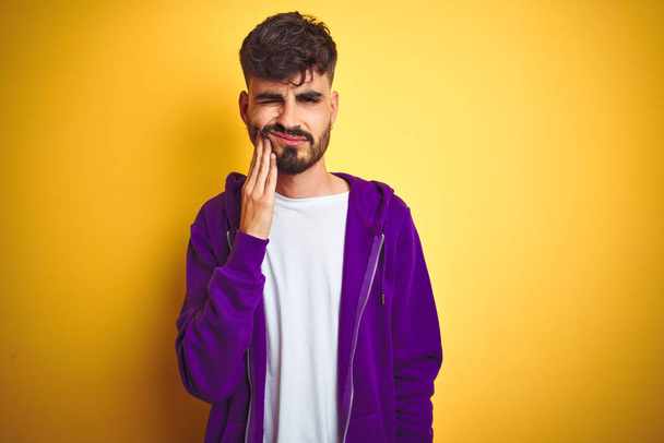 歯の痛みや歯の病気のために痛みを伴う表情で手に触れる孤立した黄色の背景の上にスポーツ紫色のスウェットシャツを着た入れ墨を持つ若い男。歯科医の概念. - 写真・画像