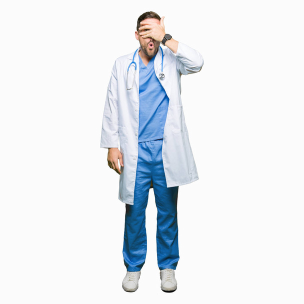 Handsome doctor man wearing medical uniform over isolated background peeking in shock covering face and eyes with hand, looking through fingers with embarrassed expression. - Photo, Image