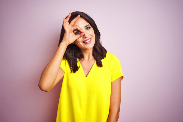 Jeune belle femme portant un t-shirt jaune debout sur fond rose isolé faisant un geste correct avec le sourire de la main, les yeux regardant à travers les doigts avec un visage heureux
. - Photo, image