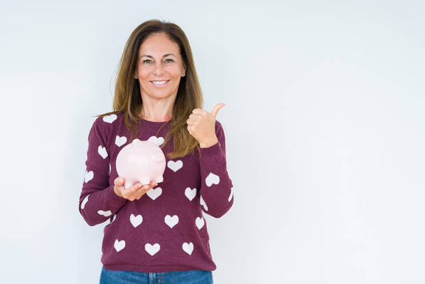 Middelbare leeftijd vrouw Holding Piggy Bank over geïsoleerde achtergrond aanwijzen en tonen met duim aan de zijkant met vrolijk gezicht glimlachend - Foto, afbeelding