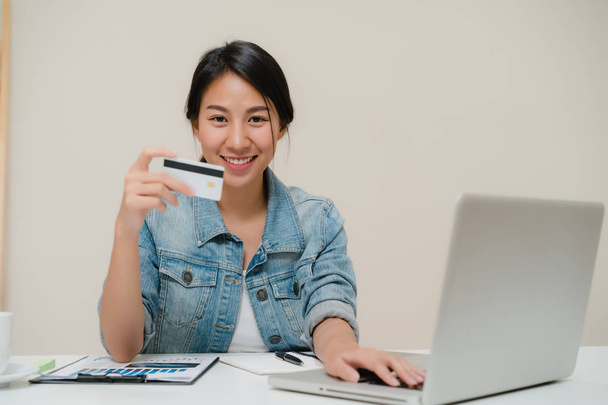 Beautiful smart business Asian woman using computer or laptop buying online shopping by credit card while wear smart casual sitting on desk in living room at home. Lifestyle woman at home concept. - Фото, изображение