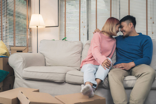 Sweet happy young Asian couple moving clothes and many objects from old home to their new home and relax on sofa in living room at their home. Lifestyle couple move new home. - 写真・画像