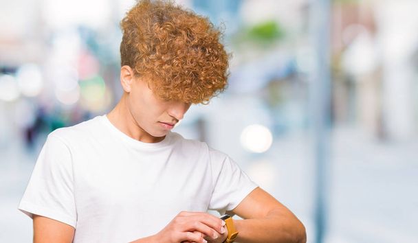 Joven hombre guapo con pelo afro con camiseta blanca casual Comprobación de la hora en el reloj de pulsera, relajado y seguro
 - Foto, Imagen