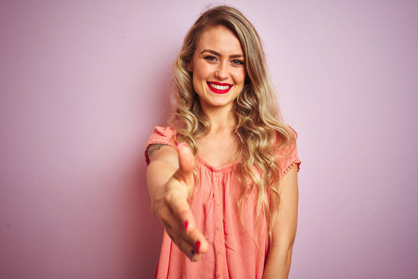 junge schöne Frau in T-Shirt vor rosa isolierten Hintergrund lächelnd freundlich mit Handschlag zur Begrüßung und Begrüßung. Erfolgreiches Geschäft. - Foto, Bild