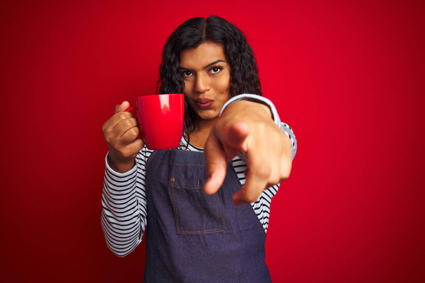 Транссексуальна транссексуальна бариста тримає чашку кави над ізольованим червоним тлом, вказуючи пальцем на камеру і до вас, знак руки, позитивний і впевнений жест спереду
 - Фото, зображення
