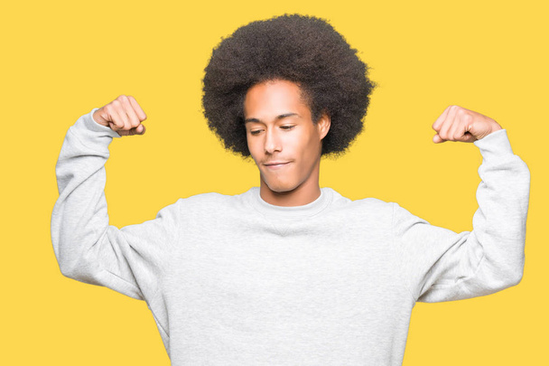 Joven afroamericano con cabello afro usando sudadera deportiva mostrando los músculos de los brazos sonriendo orgulloso. Concepto Fitness
. - Foto, Imagen