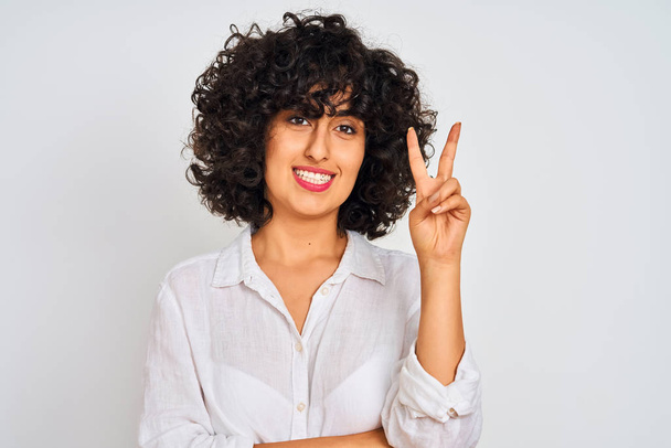 カジュアルなシャツを着た巻き毛の若いアラブ人女性は、勝利のサインをしているカメラで幸せそうな顔で微笑む。ナンバー2. - 写真・画像