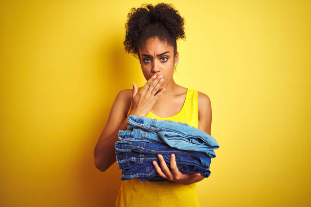 Jonge African American vrouw Holding stapel jeans over geïsoleerde gele achtergrond cover mond met de hand geschokt met schaamte voor fout, uitdrukking van angst, bang in stilte, geheime concept - Foto, afbeelding