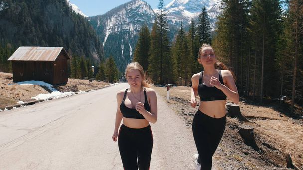 Two young athletic women in sports bra running down the road on the background of the snowy mountain - Photo, Image
