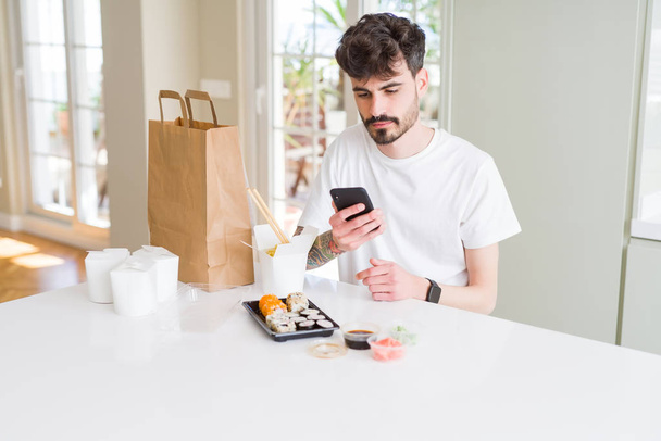Mladý muž pojídáním asijských Suši z domácích dodávek a objednávání jídla pomocí smartphone s sebevědomým výrazem na chytré tváři, který myslí vážně - Fotografie, Obrázek