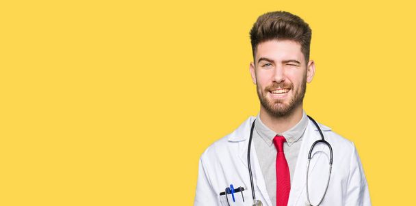 junger, gutaussehender Arzt mit medizinischem Mantel, der augenzwinkernd in die Kamera blickt, mit sexy Gesichtsausdruck, fröhlichem und glücklichem Gesicht. - Foto, Bild