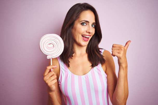 Młoda piękna kobieta jedząca słodki cukierek nad różowym białym tle wskazując i pokazując kciukiem do boku z szczęśliwą twarzą uśmiechniętą - Zdjęcie, obraz