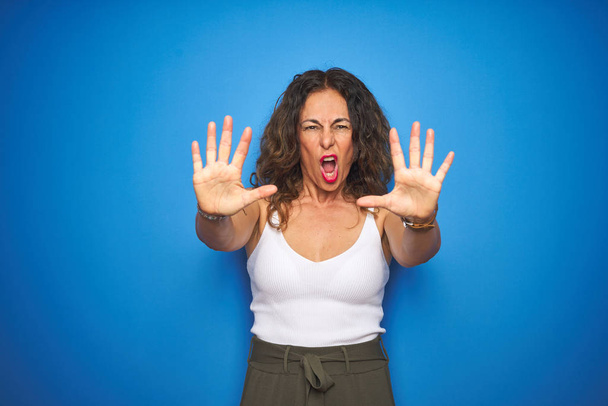 Mujer mayor de mediana edad con el pelo rizado de pie sobre fondo aislado azul haciendo gesto de stop con palmas de las manos, enojado y expresión de frustración
 - Foto, imagen