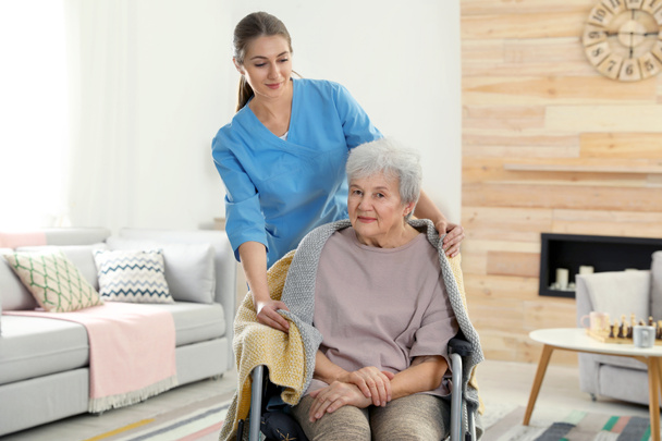 Nurse covering elderly woman in wheelchair with blanket indoors. Assisting senior people - Zdjęcie, obraz