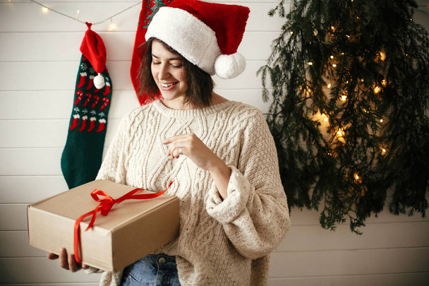 Stylish happy girl in festive sweater and santa hat opening gift - Zdjęcie, obraz