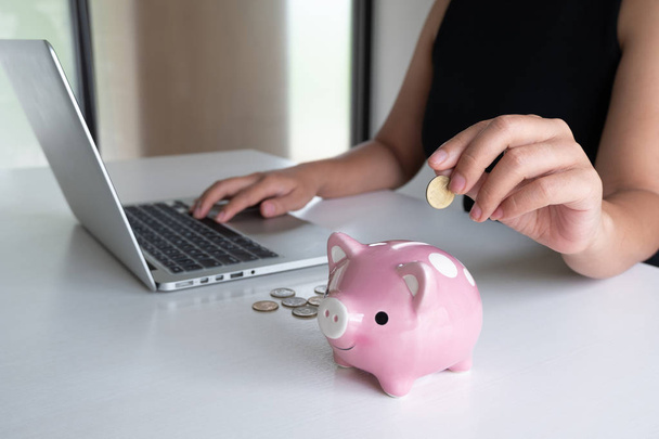 ピンクの貯金箱に金貨を入れて、オンラインマーケティングのためにラップトップを使用する女性の手は、成功へのビジネスをステップアップ、将来の計画と退職基金の概念のためのお金を節約 - 写真・画像