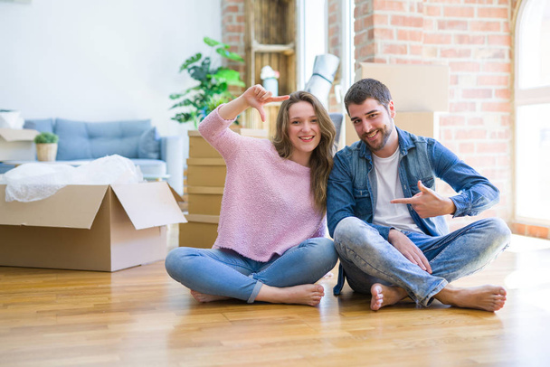 Jonge mooie paar verhuizen naar een nieuw huis zittend op de vloer glimlachend maken frame met handen en vingers met gelukkig gezicht. Creativiteit en fotografie concept. - Foto, afbeelding