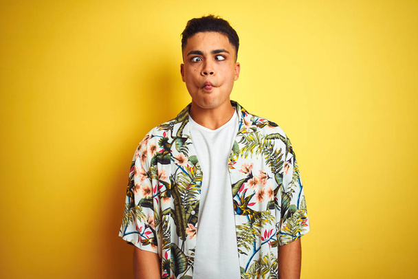 Young brazilian man on vacation wearing summer floral shirt over isolated yellow background making fish face with lips, crazy and comical gesture. Funny expression. - Photo, Image
