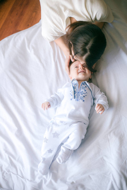 Mom's hand holds  little baby in white clothes lying on the bed at home - Foto, imagen