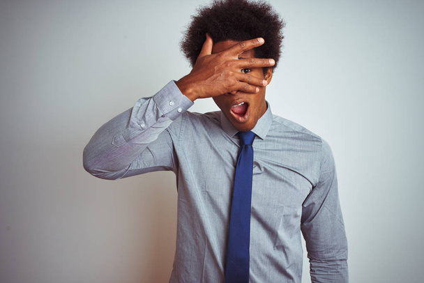 Homem de negócios americano com cabelo afro vestindo camisa e gravata sobre fundo branco isolado espreitando em choque cobrindo rosto e olhos com a mão, olhando através de dedos com expressão envergonhada
. - Foto, Imagem