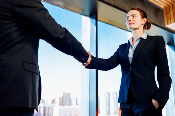 Профессиональная деловая женщина и мужчина в официальном костюме пожимают друг другу руки в помещении
. - Фото, изображение