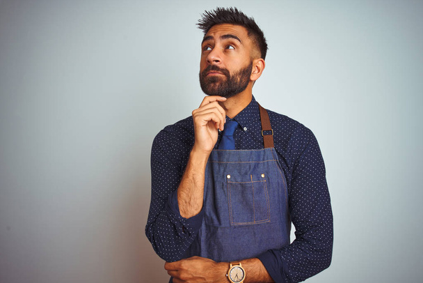 Young indian employee man wearing apron uniform standing over isolated white background with hand on chin thinking about question, pensive expression. Smiling with thoughtful face. Doubt concept. - Photo, Image