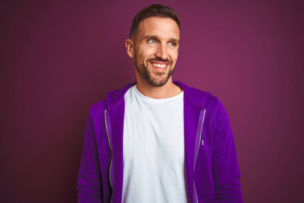 Молодой спортсмен в спортивной толстовке на фиолетовом изолированном фоне смотрит в сторону с улыбкой на лице, естественным выражением лица. Смеясь уверенно
. - Фото, изображение