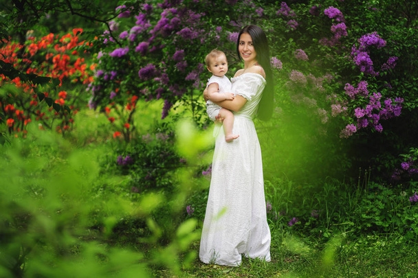 Young mother holds her little daughter in her arms among blooming trees. Lilac tree. Mothers day - Foto, afbeelding
