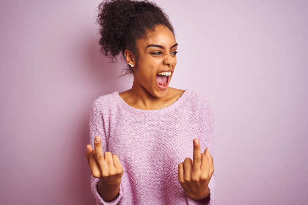 孤立したピンクの背景の上に立つ冬のセーターを着ている若いアフリカ系アメリカ人女性は、中指が悪い表情、挑発、失礼な態度をファックしている様子を見せます。興奮して叫ぶ - 写真・画像