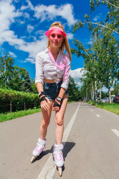 Portrait of an emotional girl in a pink cap visor and protective gloves for rollerblades and skateboarding riding on rollerblades on the road. - Φωτογραφία, εικόνα