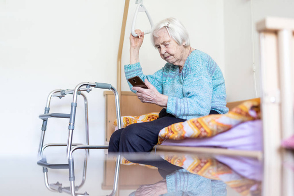 Пожилая 96-летняя женщина читает телефонные сообщения, сидя на медицинской кровати, поддерживая ее держателем
. - Фото, изображение