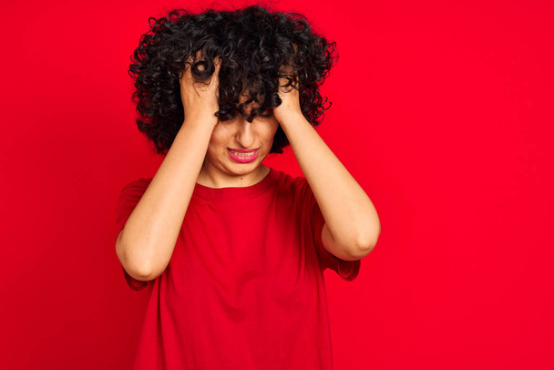 Молода арабська жінка з кучерявим волоссям носить повсякденну футболку над ізольованим червоним тлом, страждає від головного болю відчайдушно і напружено, тому що біль і мігрень. Руки на голові
. - Фото, зображення