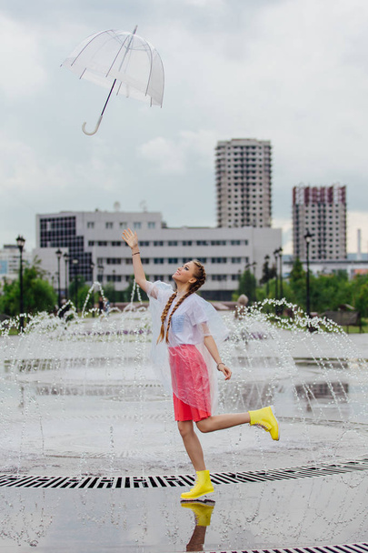 Jeune jolie fille avec deux tresses en bottes jaunes jetant et attrapant parapluie transparent près de la fontaine
. - Photo, image