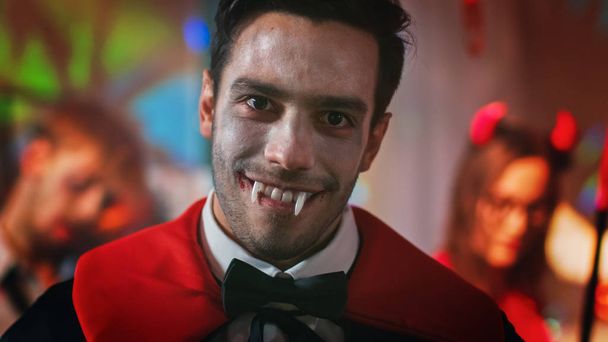 Halloween Costume Party: Portrait of Count Dracula Smiling Friendly, Shows His Deadly Bloody Fangs. In the Background Retro Lit, Decorated Room with Scary Monsters Dancing - Fotoğraf, Görsel