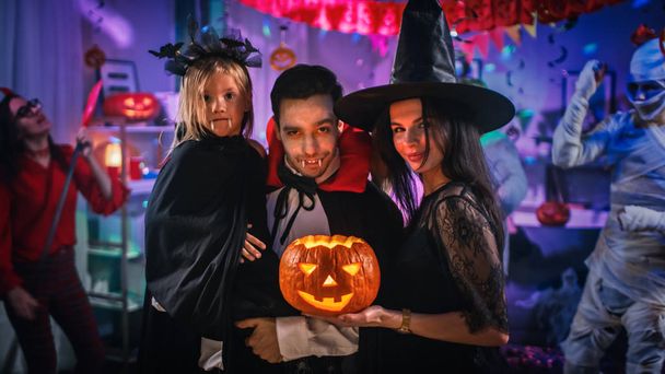 Halloween-Kostümparty: Vater Graf Dracula hält kleine Fledermausmädchen in der Hand und umarmt Hexenfrau für ein glückliches Familienporträt. Im Hintergrund Monster, die Spaß haben - Foto, Bild