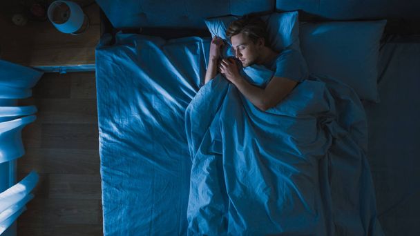 Top View of Handsome Young Man Sleeping Cozily on a Bed in His Bedroom at Night. Blue Nightly Colors with Cold Weak Lamppost Light Shining Through the Window. - Photo, Image