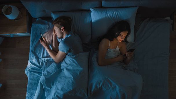 Alienated Millennial Young Couple in the Bed, Young People Turn Away From Each other Using Smartphones, Browsing Through Social Networks and Not Talking to Each Other. Top Down Camera Shot - Foto, Bild