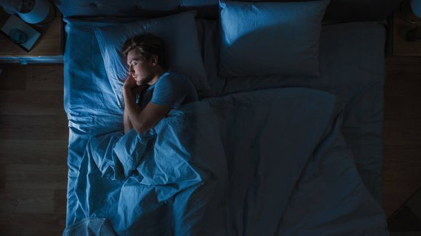 Top View of Handsome Young Man Sleeping Cozily on a Bed in His Bedroom at Night. Blue Nightly Colors with Cold Weak Lamppost Light Shining Through the Window. - Photo, Image