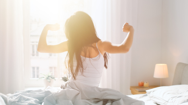 Beautiful Brunette is Waking up in the Morning, Stretches in the Bed, Sun Shines on Her From the Big Window. Happy Young Girl Greets New Day with Warm Sunlight Flare. - Photo, Image