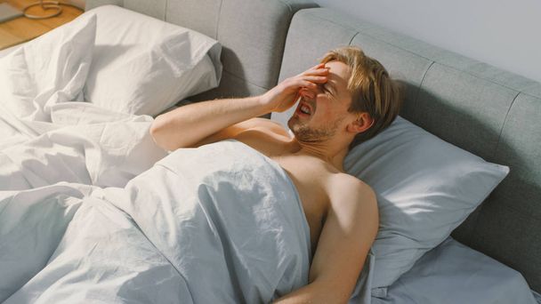 Unhappy Young Man Wakes Up in the Morning Because of a Sun. Tired with the Pain in Head, Grimacing, Probably Hangover. Bad Morning for a Fit Guy. - Foto, Bild