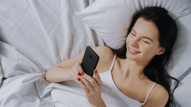 Portrait of Beautiful Brunette Using Touchscreen Smartphone in Her Bed, Chatting with Friends, Using Social Media, Posting Pictures, Sharing Thoughts. Top Down Shot. - Photo, image