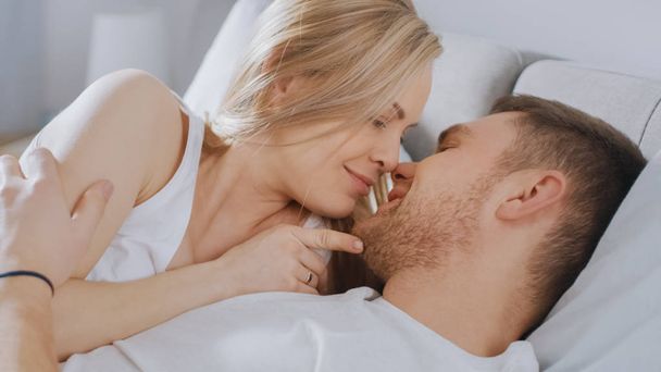 Ευτυχισμένο νεαρό ζευγάρι αγκαλιάζοντας μαζί στο κρεβάτι, φιλιά και χαμόγελα. Όμορφη ξανθιά γυναίκα και της ταιριάζει στοργικό σύντροφο βιώνει τρυφερό και στοργικό στιγμές του ζώντας μαζί. - Φωτογραφία, εικόνα