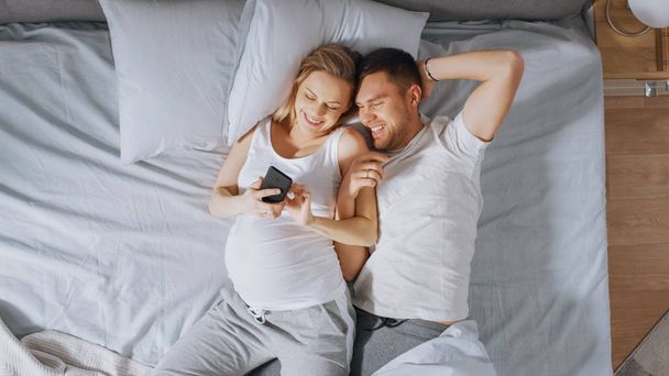 Loving Young Couple Spending Morning in Bed, Pregnant Young Woman Shows Her Partner Something on a Touchscreen Smartphone. Searching for Baby Clothes, Browsing Through Social Media, Posting Pictures. - Zdjęcie, obraz