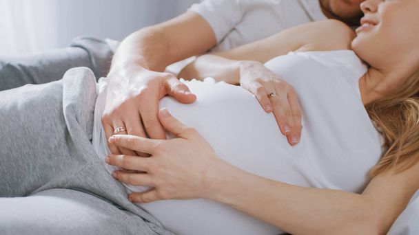 Happy Young Couple Cuddling Together in the Bed, Young Woman is Pregnant and Loving Husband Touches and Caresses Her Belly Tenderly. Camera Focus on the Belly. - Foto, afbeelding